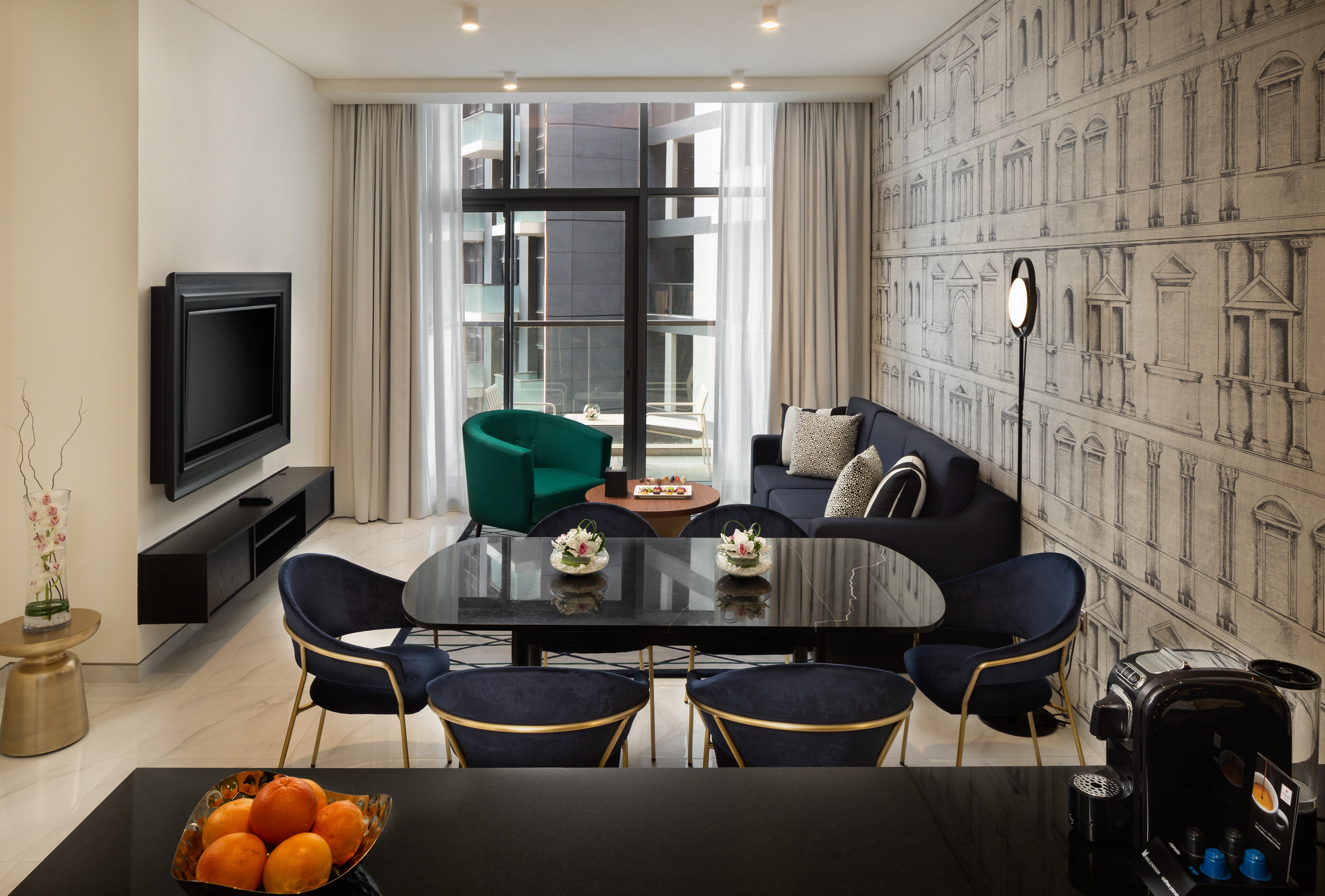 Aparthotel Millennium Atria Business Bay Dubaj Exteriér fotografie The photo shows a modern living space that includes a cozy seating area and dining setup. On the left, there is a large television mounted on the wall. The living room features a blue sofa and a green accent chair, creating a stylish contrast. A smal