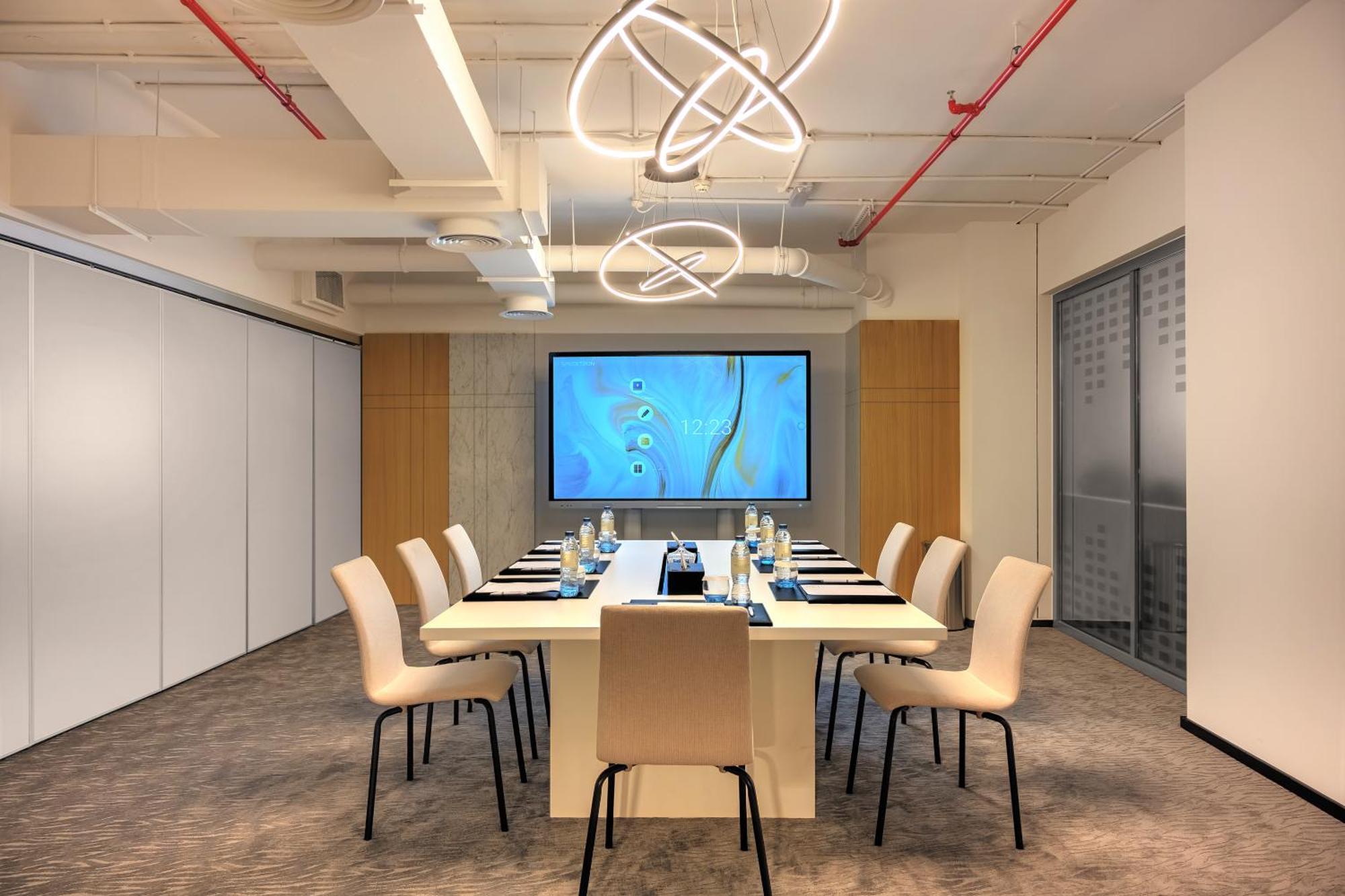 Aparthotel Millennium Atria Business Bay Dubaj Exteriér fotografie The photo shows a modern conference room designed for meetings. It features a long white table surrounded by beige chairs. At the end of the table, there is a large screen displaying a blue abstract design. The room is well-lit with contemporary ligh