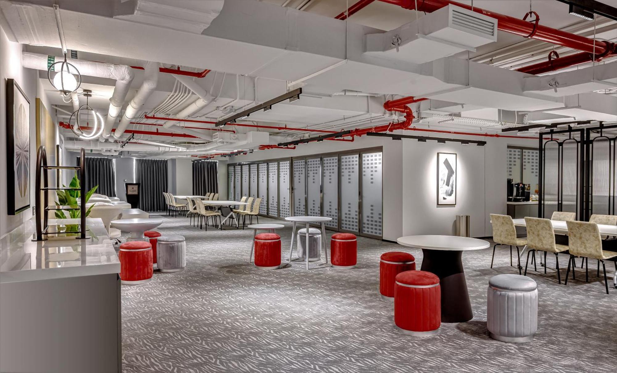 Aparthotel Millennium Atria Business Bay Dubaj Exteriér fotografie The photo shows a modern interior space, likely an office or communal area. The flooring is covered with a textured carpet, and the ceiling features exposed pipes painted red. 

There are several seating options, including small round tables with sto