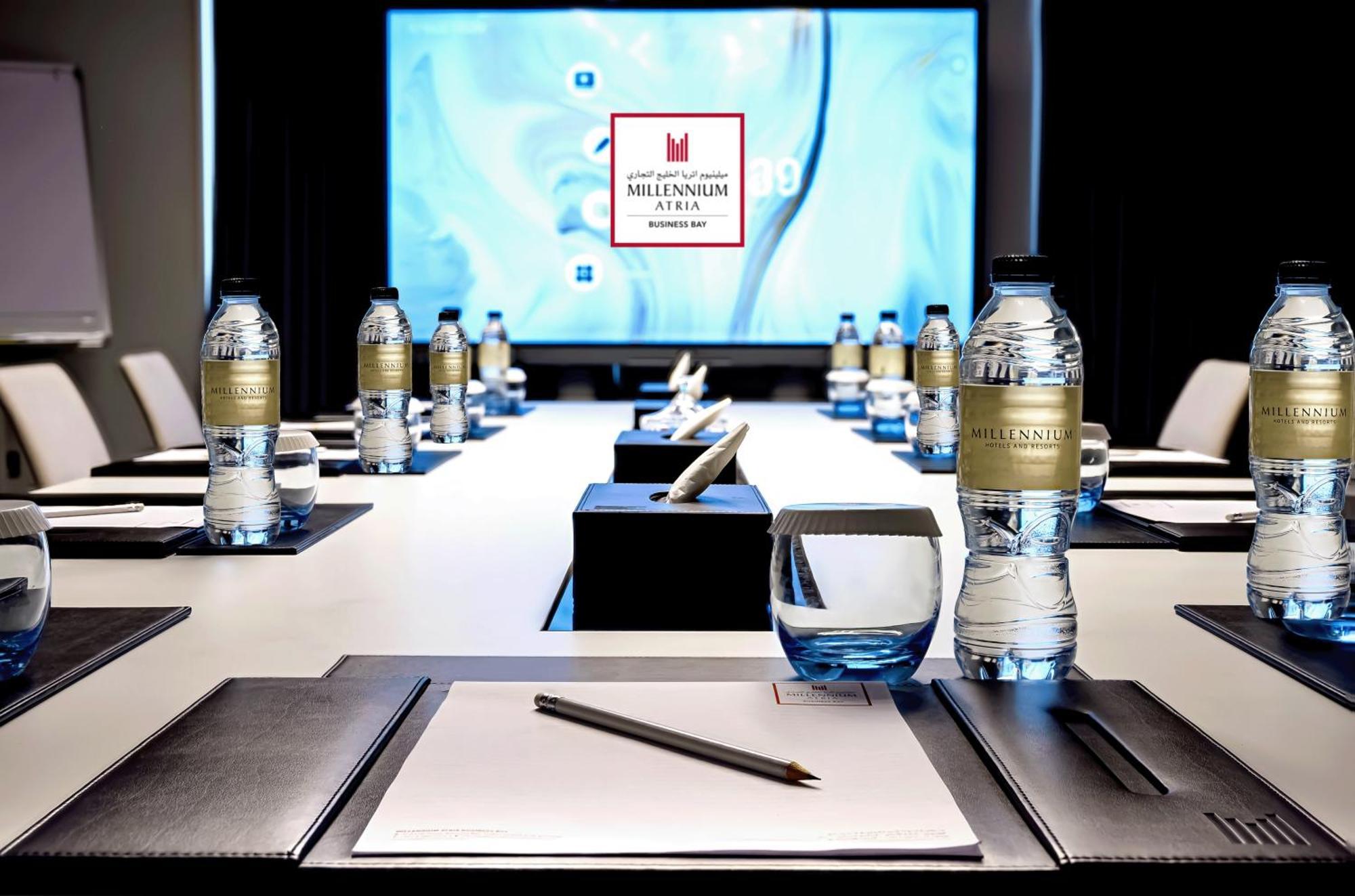 Aparthotel Millennium Atria Business Bay Dubaj Exteriér fotografie The photo shows a modern conference room setup. A long table is laid out with neatly arranged stationery, including notebooks and pens, at each seat. Several bottles of water are placed on the table. In the background, a large screen is displayed, li