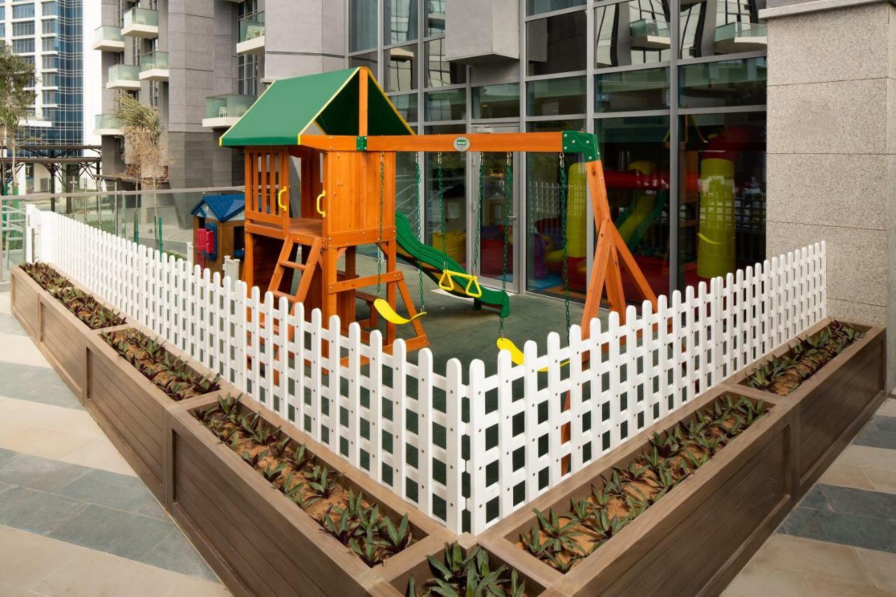 Aparthotel Millennium Atria Business Bay Dubaj Exteriér fotografie The image shows a children's playground area, which is enclosed by a white picket fence. The playground features a wooden play structure with a slide, swings, and climbing elements. Surrounding the play area are planters with greenery, and the backdr
