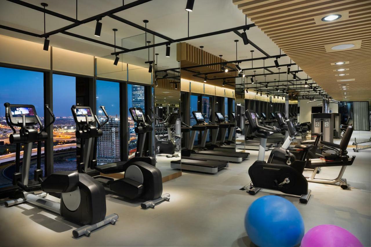 Aparthotel Millennium Atria Business Bay Dubaj Exteriér fotografie The photo shows a modern gym with various types of exercise equipment available. There are several treadmills and elliptical machines arranged in rows, with large windows in the background providing a view of the city. The lighting is bright and cont