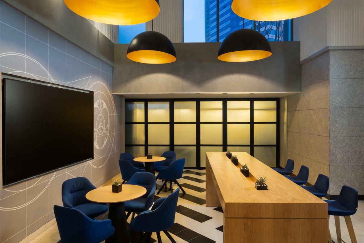 Aparthotel Millennium Atria Business Bay Dubaj Exteriér fotografie The photo shows a modern interior space, likely a lounge or meeting area. It features a long wooden table with some small plants on it, accompanied by several blue, circular chairs arranged around it. There is a large flat-screen television mounted o