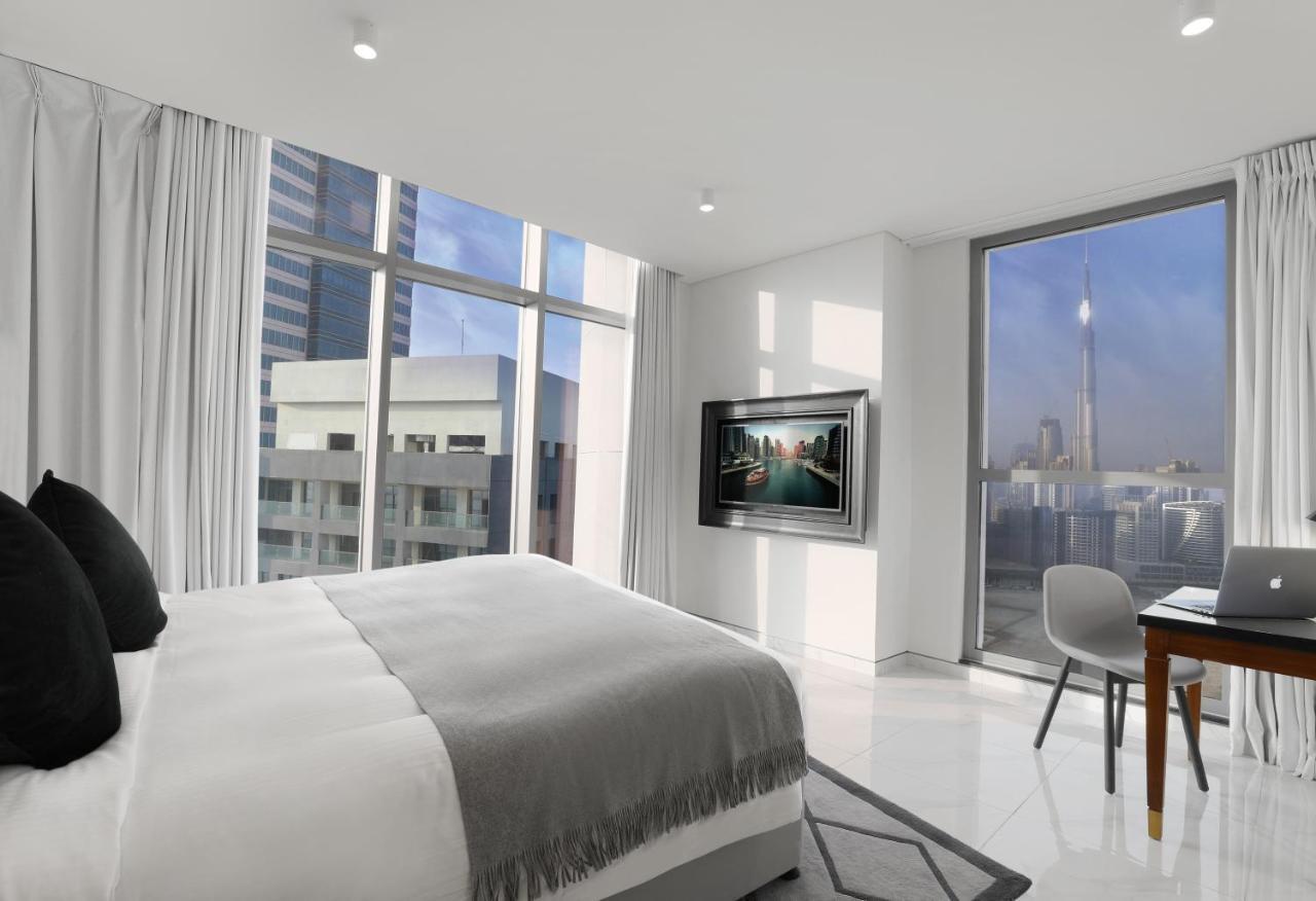 Aparthotel Millennium Atria Business Bay Dubaj Exteriér fotografie The photo depicts a modern hotel room that features a large bed with a gray throw blanket. There are large windows allowing natural light to fill the space, showcasing a city skyline view. A flat-screen television is mounted on the wall opposite the 