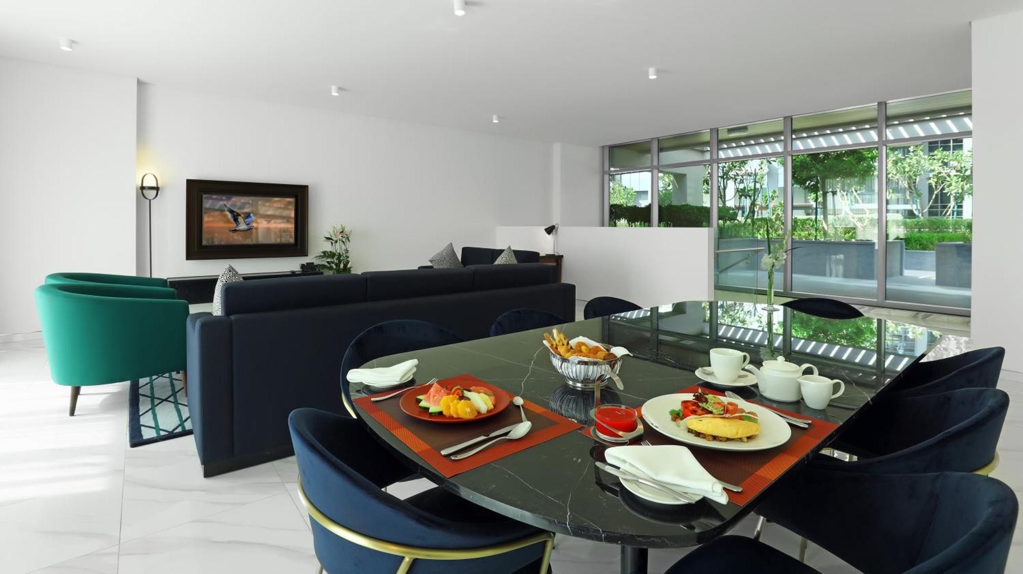 Aparthotel Millennium Atria Business Bay Dubaj Exteriér fotografie The photo depicts a modern living space with a dining area and a seating area. In the foreground, there is a round dining table set with plates of food, including eggs, toast, and a small bowl of fruit, accompanied by cups and napkins. The table is c